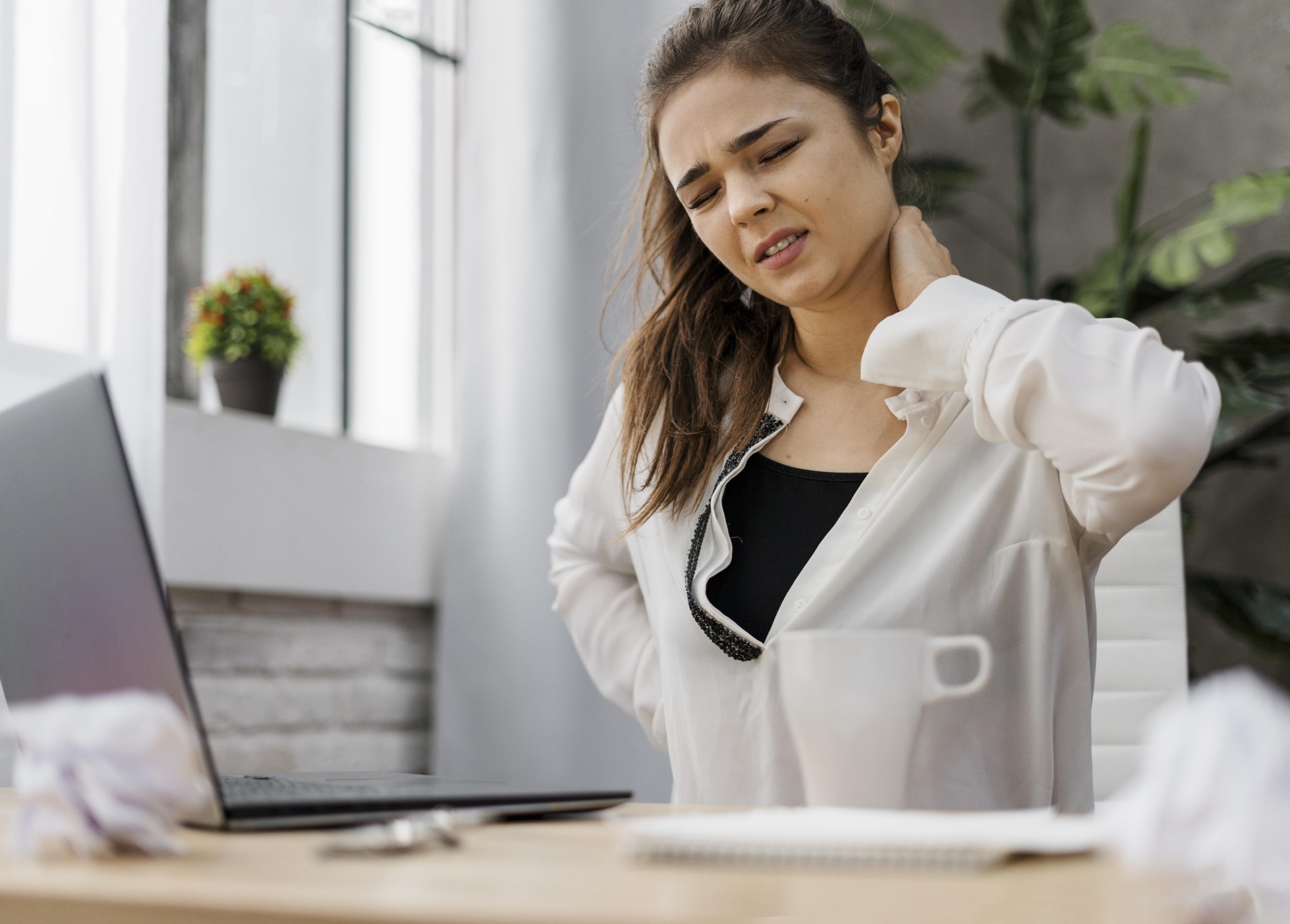 A woman sitting at a desk, looking distressed and holding her neck and shoulder in pain.